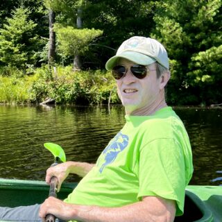 man wearing yellow shirt holding a paddle in a green kayak on a pond with trees and pond in the background